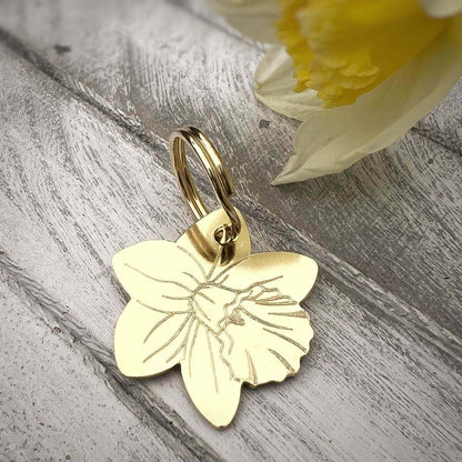 Gold daffodile dog tag on a wooden surface with a yellow flower in the background