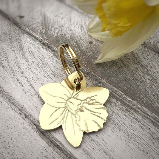 Gold daffodile dog tag on a wooden surface with a yellow flower in the background