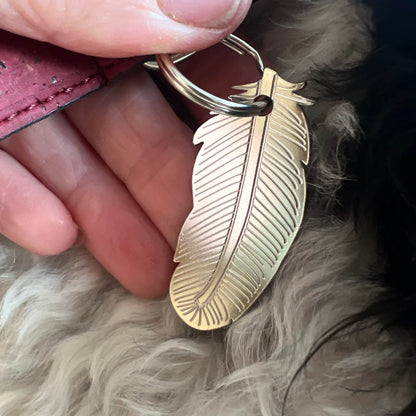 A close-up of a hand holding a silver-toned feather-shaped pet ID tag attached to a key ring, with fluffy light-colored fur in the background.
