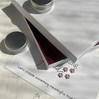 Open white box with red tissue paper on a white surface, accompanied by a card with text and paw prints and metal tins. 