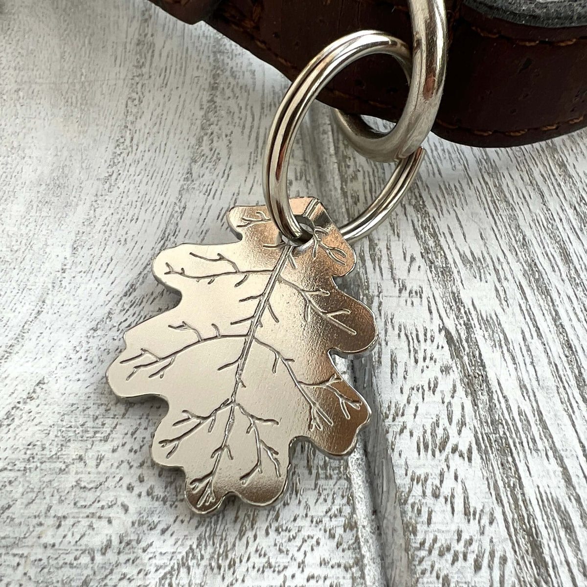 Silver colour oak leaf-shaped tag attached to a silver split ring and dark brown leather collar, set against a white-washed wood background.