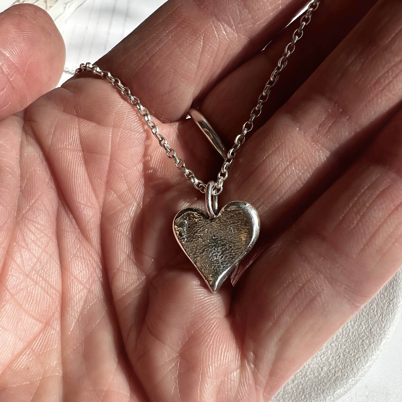 Silver heart-shaped pendant on a chain held in a hand against a neutral background