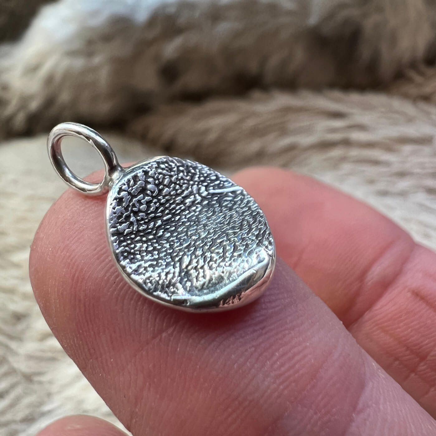 Silver paw print textured round pendant held on fingers against a blurred fur background