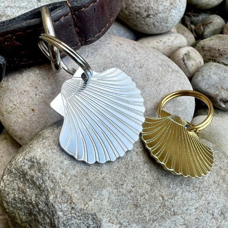 Two seashell shaped dog ID tags, one silver-toned and one gold-toned, displayed on a large gray rock next to a dark brown leather collar.