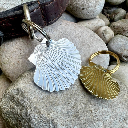 Two seashell shaped dog ID tags, one silver-toned and one gold-toned, displayed on a large gray rock next to a dark brown leather collar.