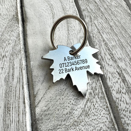 A personalized metal pet ID tag shaped like a maple leaf, engraved with a name and address, attached to a silver split ring on a rustic whitewashed wood surface.