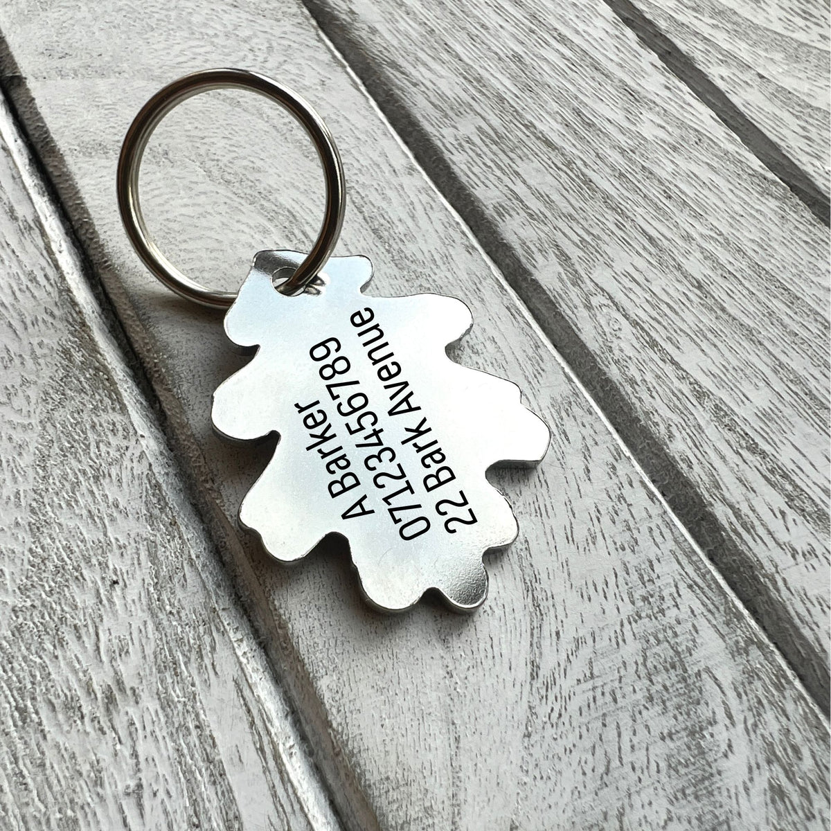 A silver metal dog tag with a custom engraved oak leaf-shaped tag on a weathered gray wooden background.