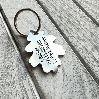 A silver metal dog tag with a custom engraved oak leaf-shaped tag on a weathered gray wooden background.