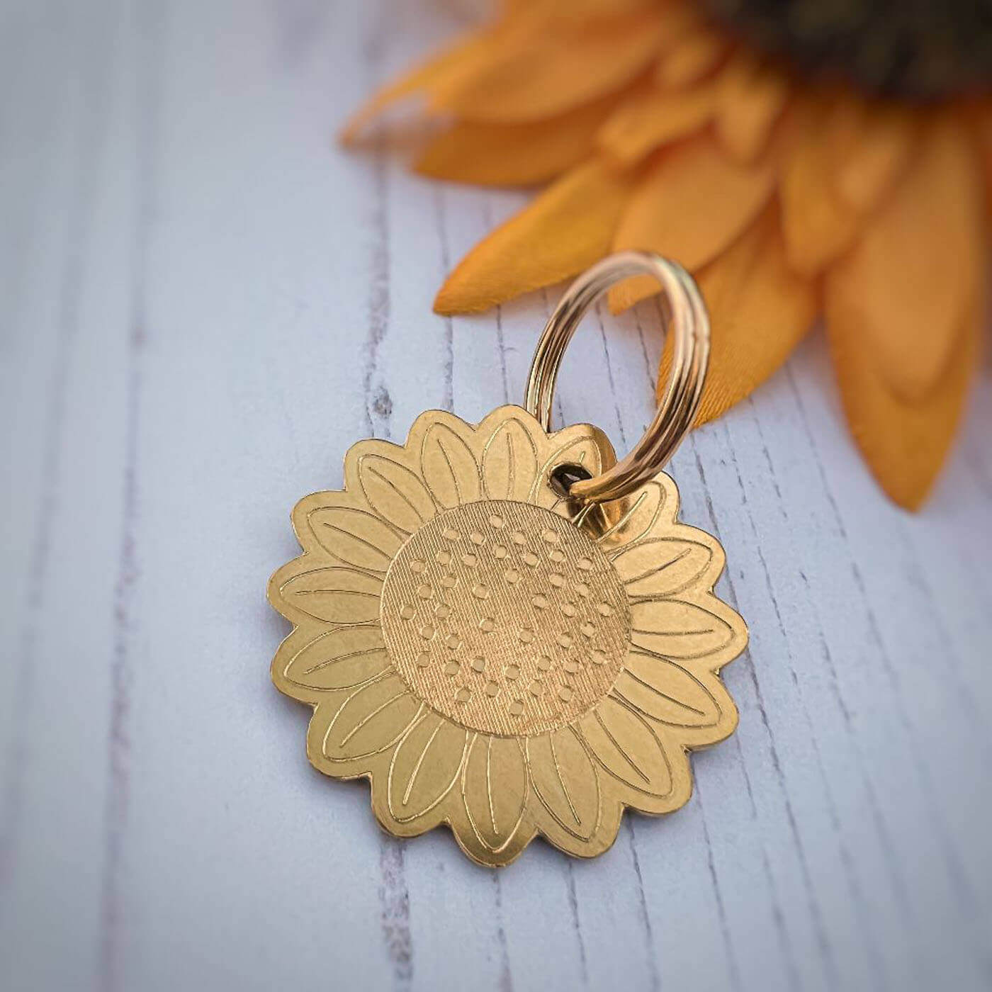 Gold sunflower-shaped dog ID tag on a light wooden surface with a sunflower in the background.